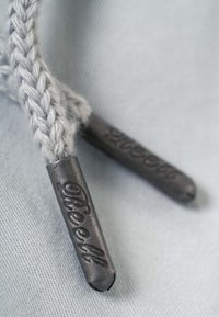 Primer plano de puntas de cordones trenzados beige con terminales de metal texturizado con la palabra "Reell" grabada sobre fondo de tela clara.