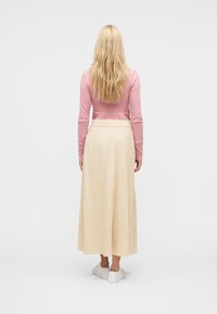 Γυναίκα με μακριά ξανθά μαλλιά που φοράει ροζ μπλούζα με μακριά μανίκια, μπεζ maxi φούστα και λευκά sneakers, στέκεται γυρισμένη ανάποδα σε απλό φόντο.