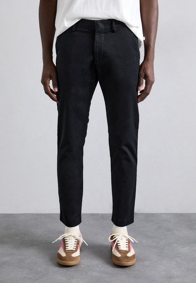 Schwarze Baumwollhosen mit schlankem Schnitt, ausgestattet mit Seitentaschen und flachem Vorderteil. Kombiniert mit mehrfarbigen Sneakers und weißen Socken.