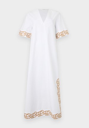 Vrouw staand, gekleed in een witte enkellange jurk met gouden geborduurde biezen aan mouwen, zoom en zijkanten, gecombineerd met bruine platte sandalen.