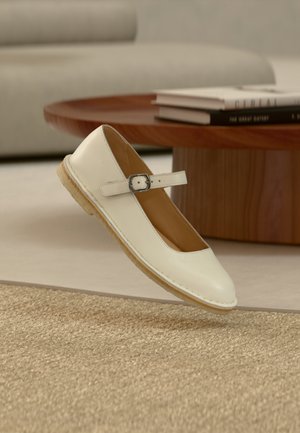 Cream-colored Mary Jane shoe with buckle strap and beige sole floating above woven rug, wooden table and books in background.
