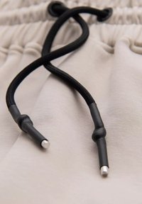 Béžová látka s texturovaným povrchem, s černými šňůrkami. Konce šňůrek jsou zakončeny kovovými akcenty.