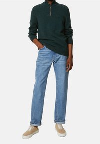 Donkergroene zip-necksweater, blauwe relaxte spijkerbroek en beige sneakers met witte zolen. Sweater heeft geribde manchetten en zoom.