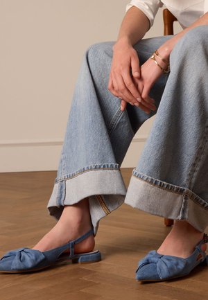 Des chaussures slip-on en textile bleu avec un détail noué au niveau des orteils, portées avec un jean en denim à jambes larges présentant un ourlet effiloché et une couture contrastante.