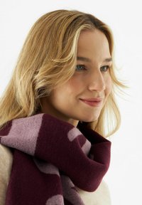 Gebreide sjaal in diep bordeauxrood, met een patroon en lichtpaarse accenten. Zachte textuur, nonchalant om de nek gedrapeerd.