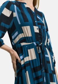 Button-up dress in teal, navy, and beige with geometric pattern. Features a tied waist, long sleeves, and soft, flowing fabric texture.