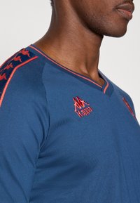 Niebieska koszula z długim rękawem i dekoltem w serek, zdobiona czerwoną haftem oraz bocznymi akcentami z powtarzającym się wzorem logo. Miękki, elastyczny materiał.