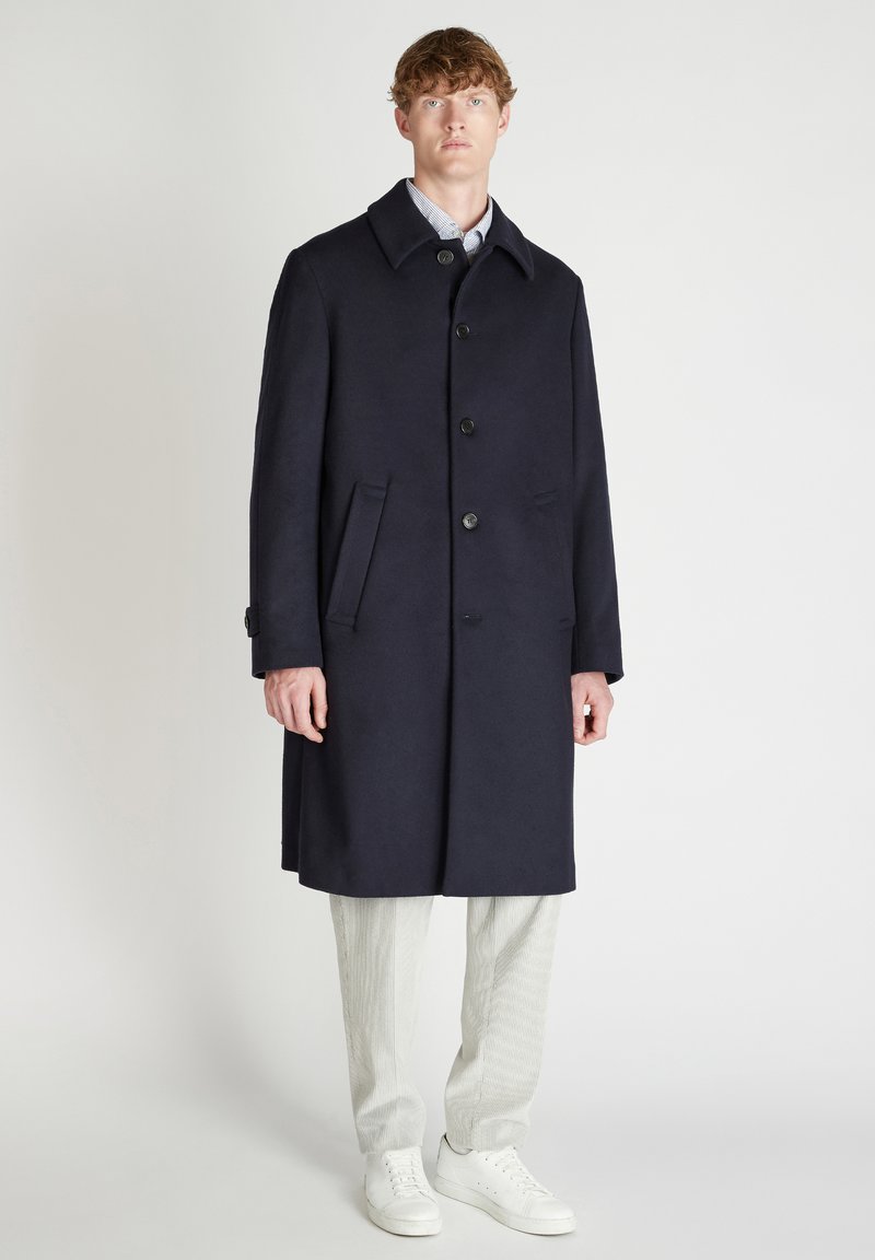 Giovane uomo in piedi, che indossa un soprabito blu scuro con bottoni, pantaloni chiari e sneakers bianche su uno sfondo bianco e semplice.