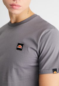 Graues Baumwoll-T-Shirt mit rundem Halsausschnitt. Verfügt über einen schwarz-roten Logo-Aufnäher auf der Brust und einen kleineren Aufnäher am Ärmel.