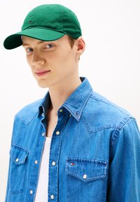 Căciulă de baseball verde din material textil, purtată împreună cu o cămașă din denim albastru cu butoane tip snap și două buzunare pe piept. Tricou alb dedesubt.