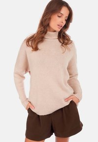 Pull en tricot doux beige avec un col montant, coupe ample et poignets côtelés. Présente un motif texturé et des poches latérales, associé à un short marron.
