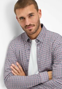 Camicia a scacchi in rosso, navy e bianco, indossata sopra una maglietta bianca. La camicia presenta un colletto classico e polsi con bottoni.
