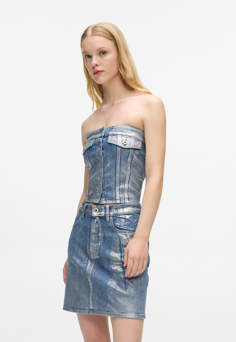 Γυναίκα φορώντας ένα λαμπερό μεταλλικό μπλε strapless τζιν τοπ και ταιριαστή τζιν φούστα, που στέκεται μπροστά σε ένα απλό λευκ�ό φόντο.