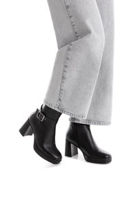 Botas de cuero negras hasta el tobillo con un tacón grueso, punta redondeada y detalle de hebilla en el lateral. Llevadas con unos vaqueros de denim gris claro de corte holgado.