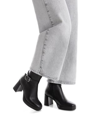 Botas de cuero negras hasta el tobillo con un tacón grueso, punta redondeada y detalle de hebilla en el lateral. Llevadas con unos vaqueros de denim gris claro de corte holgado.