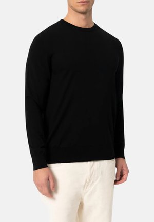 Uomo che indossa un maglione nero semplice a maniche lunghe con scollo a girocollo e pantaloni bianco sporco, in piedi su uno sfondo chiaro.