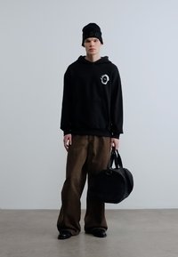 Μαύρο φούτερ με κουκούλα και λογότυπο, φαρδιά καφέ παντελόνια, μαύρο σκουφάκι και μαύρη στρογγυλή τσάντα, φορώντας από ανδρική φιγούρα σε μινιμαλιστικό περιβάλλον.