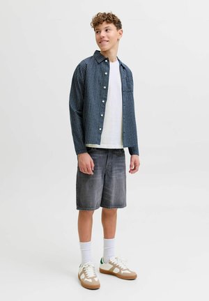 Adolescent portant une chemise à carreaux bleue boutonnée sur un t-shirt blanc, un short en denim gris, des chaussettes blanches et des baskets beige blanc, debout et souriant.