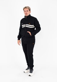 Giacca nera con zip e righe bianche, tasche laterali e logo sul petto, abbinata a pantaloni jogger neri e sneakers bianche.