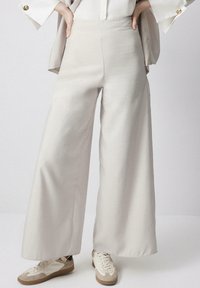 Ljusbeige vida byxor i mjukt tyg med en slät textur, med hög midja och sidfickor, tillsammans med avslappnade sneakers.