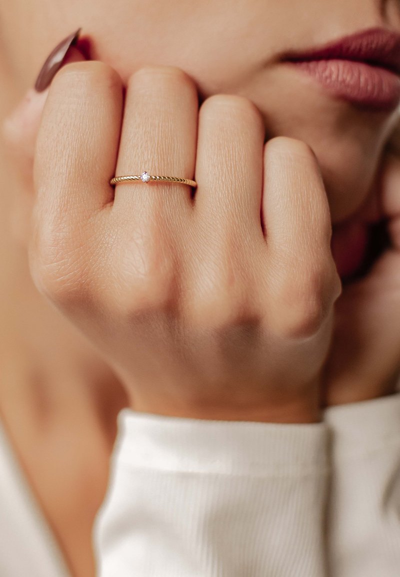 Anello d'oro con un design spiralato, caratterizzato da una piccola pietra preziosa trasparente, indossato su una mano con pelle leggermente abbronzata. Lo sfondo è delicatamente sfocato.