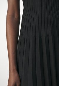 Gros plan d’un bras à la peau foncée à côté d’une robe ou jupe noire côtelée et plissée.