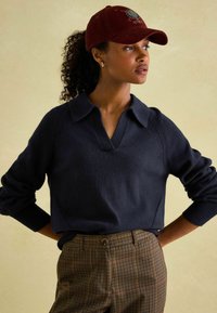 Dunkelblauer Pullover mit kragenähnlichem Ausschnitt und strukturierten Schulterdetails, kombiniert mit braunen karrierten Hosen. Burgunderfarbener Hut mit gesticktem Design.