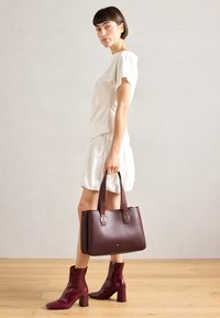 Sac en cuir marron avec doubles poignées, design structuré et accents en matériel doré, associé à une tenue blanche et des bottes à talons marron.