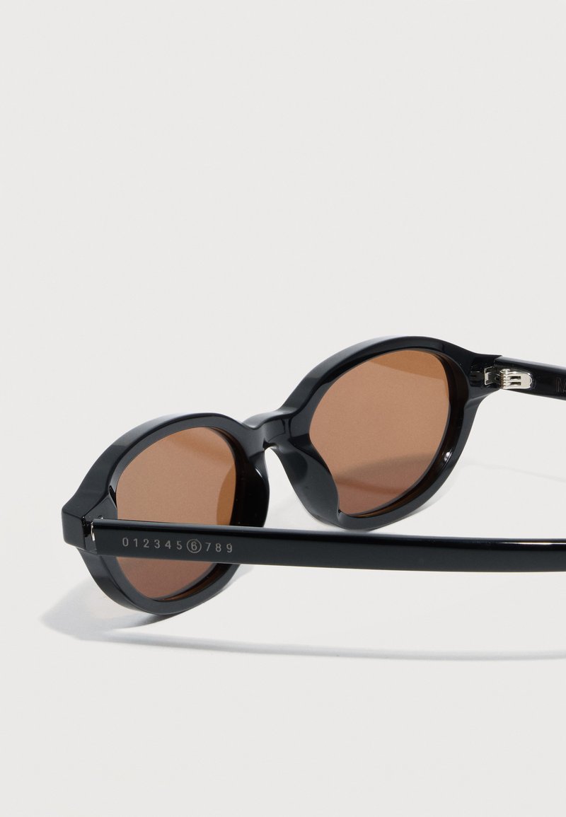 Black round-frame sunglasses with brown tinted lenses resting on a light surface, showing numeric detail on one temple arm.