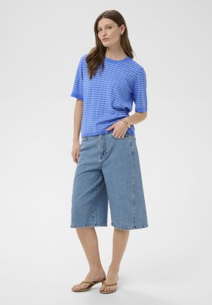 Femme portant un pull à manches courtes rayé bleu, un short en jean longueur genou et des sandales à talons beiges, se tenant debout avec une main dans la poche.