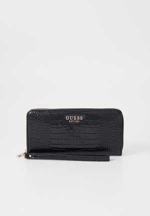 Cartera de piel con grabado de cocodrilo negro, con un logo dorado "GUESS EST 1981", cierre con cremallera y correa desmontable para facilitar su transporte.