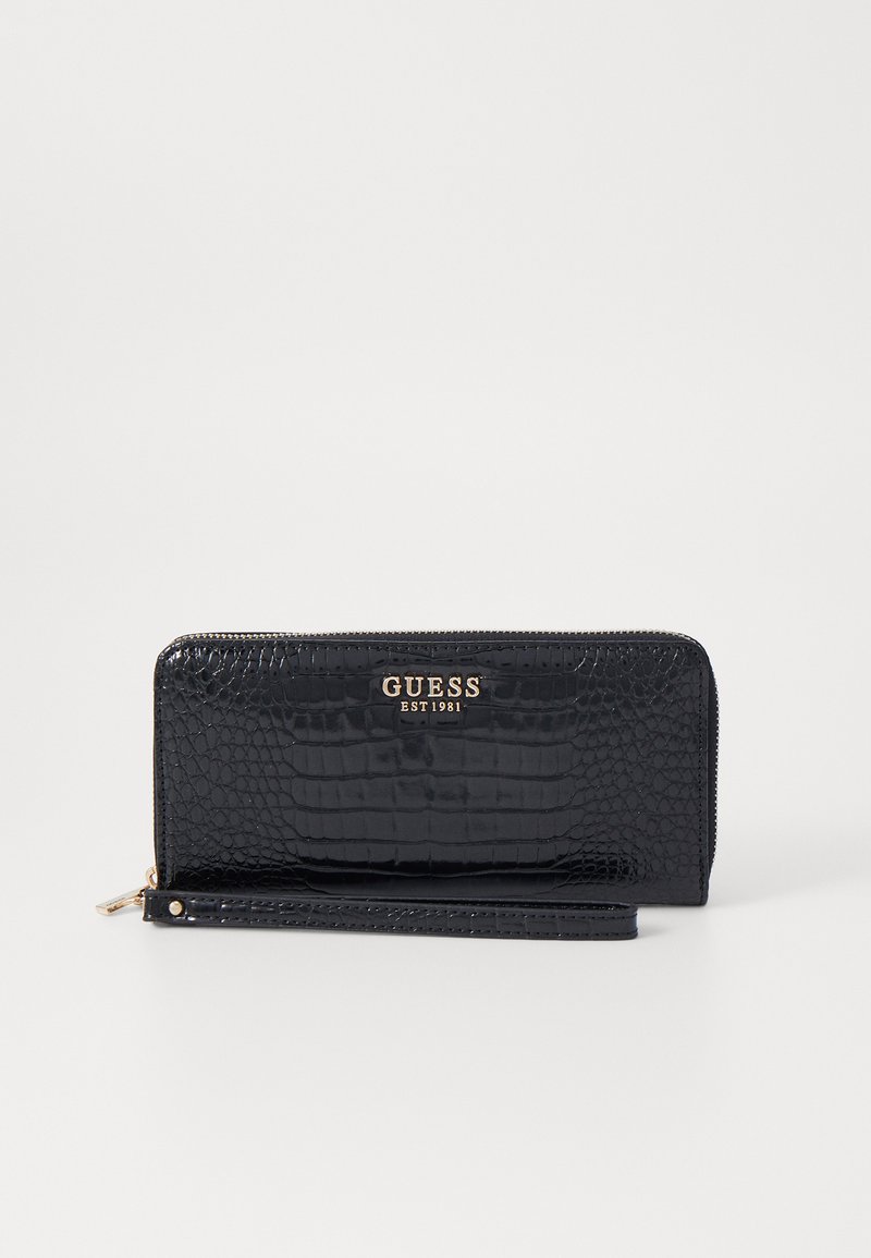 Cartera de piel con grabado de cocodrilo negro, con un logo dorado "GUESS EST 1981", cierre con cremallera y correa desmontable para facilitar su transporte.
