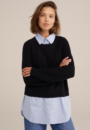 Un pull noir en tricot avec des détails côtes sur une chemise à boutons bleu clair et blanche à rayures, associé à un jean en denim.