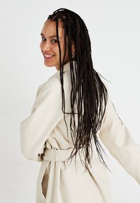 Beige színű trench coat, derékra kötött övvel, sima textúrával és hosszú ujjakkal, fonott hajjal viselve, egy sima háttér előtt.