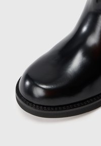 Bota de tobillo de cuero negro con acabado brillante, puntera redondeada y suela negra cosida en contraste. Textura suave con un diseño elegante.