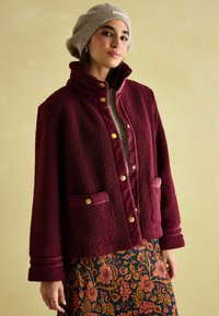 Burgundia fliisjakker, millel on nööbid ja kaks esist meest, silmapaistva pehme tekstuuriga ja kontrastse sametäärisega. Kombineeritud mustrilise seelikuga.