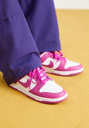 Nike sneakers met een witte bovenkant en roze accenten, een ronde neus en roze veters. Gecombineerd met losse, donkerpaarse broek.