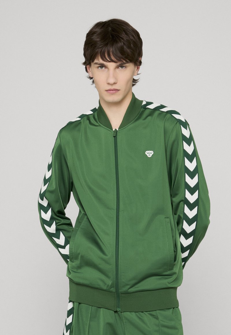 Veste zippée verte en tissu lisse avec des rayures chevrons blanches sur les manches et un petit emblème sur la poitrine, dotée d'un col et de poignets en tricot.