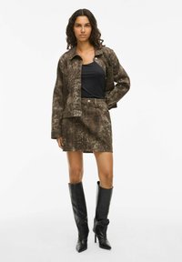 Giacca in stampa pitone marrone sopra una maglia nera, abbinata a una gonna coordinata e stivali neri al ginocchio. Tessuto testurizzato con un design strutturato.