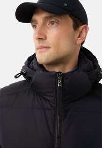 Zwarte pufferjack met een hoge kraag en ritssluiting. Bevat aantrekkoorden en een textuurstof, vergezeld van een marineblauwe pet.