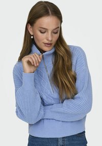 Lyseblå strikkegenser med høy hals og glidelås, med teksturerte kabel- og ribbestrikkede mønstre, og voluminøse ermer.