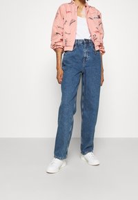 Růžová bomber bunda s ručně psanými vzory, bílé tílko, modré džíny s vysokým pasem a bílé tenisky. Ležérní outfit na jednobarevném pozadí.