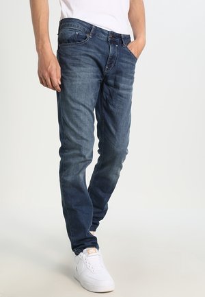 Mand iført mørkeblå slim-fit jeans og hvide sneakers, stående med den ene hånd i lommen mod en ensfarvet baggrund.