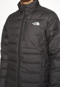 Svart vadderad jacka med hög krage, dragkedja, quiltat mönster och en logotyp av The North Face på vänster bröst.