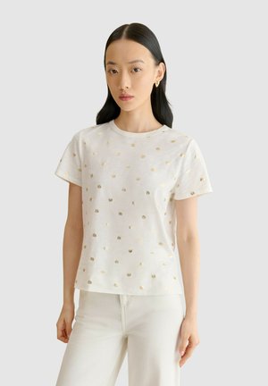 Mujer con cabello largo y negro, vestida con una camisa blanca de manga corta con pequeños patrones de sol dorados y pantalones blancos, de pie frente a un fondo liso.
