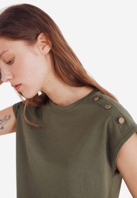 Oliven grøn t-shirt med korte ærmer i blød bomuld, med rund halsudskæring og guldknapdetaljer på skulderen. Glat tekstur.