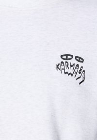 Sweatshirt blanc avec impression graphique noire de mains stylisées et du texte "KARMA" sur la partie gauche de la poitrine. Tissu doux et texturé.