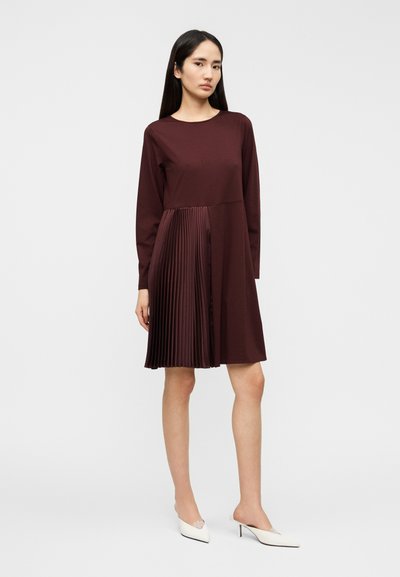 Μπορντό μακρυμάνικο φόρεμα με εφαρμοστή κορυφή και πλισέ φούστα. Ομαλή υφή, μήκος ως το γόνατο και στρογγυλή λαιμόκοψη.