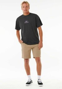 T-shirt grigio scuro a maniche corte con logo bianco, abbinato a pantaloni corti beige e sneakers nere con suole bianche e lacci a fantasia.