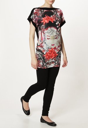 Person iført en kortærmet tunika med rødt blomster- og geishaansigtsprint, sorte leggings og sorte balletsko, stående med det ene ben løftet.